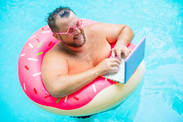 Усміхнений товстий смішний чоловік в рожевому надувному колі в рожевих окулярах працює на ноутбуці в басейні — стокове фото