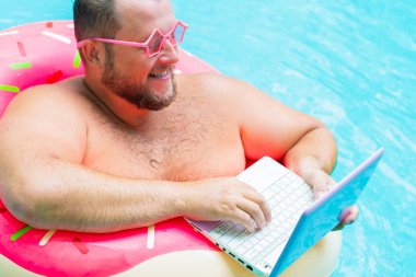 pembe gözlük pembe şişme daire içinde gülümseyen Şişman komik adam bir yüzme havuzunda bir dizüstü bilgisayar üzerinde çalışır
