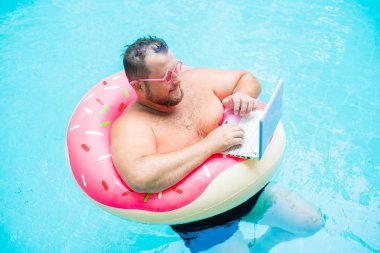 pembe gözlük pembe şişme daire içinde ciddi Şişman komik adam bir yüzme havuzunda bir dizüstü bilgisayar üzerinde çalışır