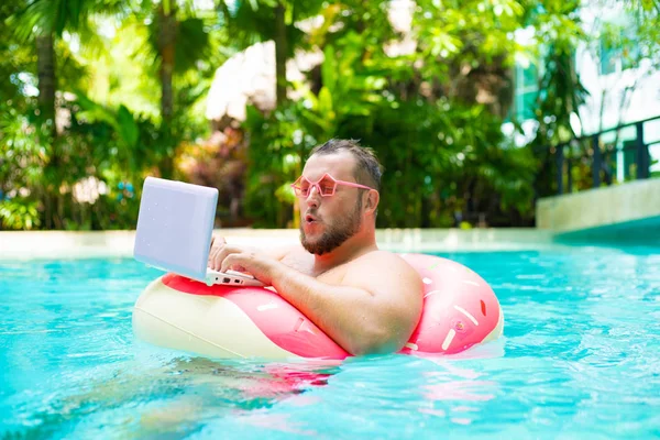 Здивований товстий смішний чоловік в рожевому надувному колі в рожевих окулярах працює на ноутбуці в басейні — стокове фото