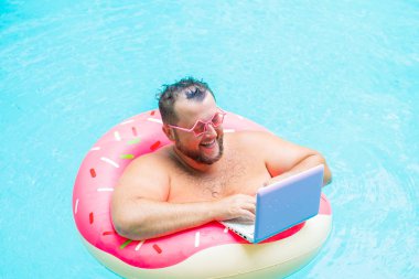 pembe gözlük pembe şişme daire içinde gülümseyen Şişman komik adam bir yüzme havuzunda bir dizüstü bilgisayar üzerinde çalışır