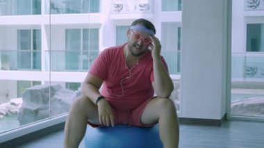 Pembe gözlük ve pembe bir t-shirt komik şişman erkek bir kız tasvir spor salonunda uygun bir top üzerinde meşgul. Bir ucube adam spor salonunda topu bir baş ağrısı var.