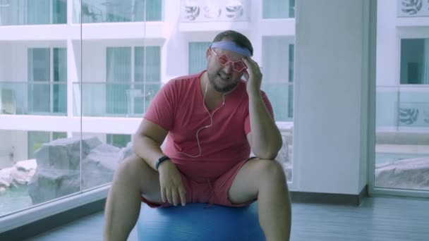 Αστείο λίπος αρσενικό σε ροζ γυαλιά και σε ένα ροζ t-shirt ασχολούνται με μια μπάλα Fit στο γυμναστήριο που απεικονίζει ένα κορίτσι. Ένας άνθρωπος φρικιό έχει πονοκέφαλο στην μπάλα στο γυμναστήριο.. — Αρχείο Βίντεο