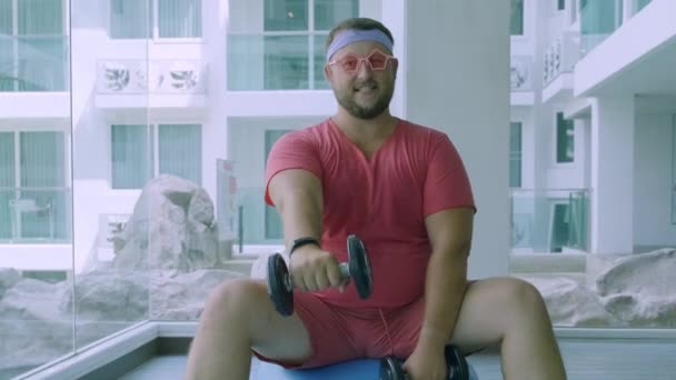 Funny fat male in pink glasses and in a pink t-shirt is engaged with dumbbells on a fit ball in the gym depicting a girl — Stock Video