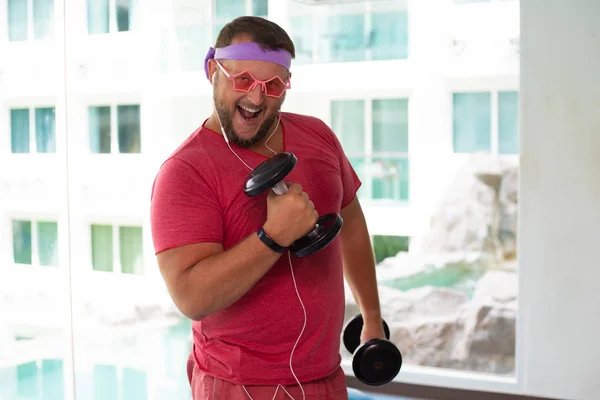 Grappige dikke man in roze bril en in een roze T-shirt bezig met dumbbells in de sportschool portretteren van een meisje. man luisteren naar muziek — Stockfoto