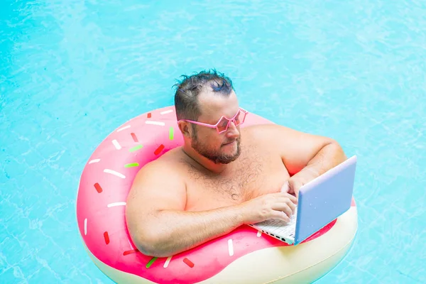 Серйозна товста смішна людина в рожевому надувному колі в рожевих окулярах працює на ноутбуці в басейні — стокове фото