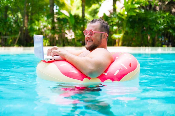 Glimlachend vet grappig man in roze opblaasbare cirkel in roze bril werkt op een laptop in een zwembad — Stockfoto