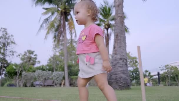 Klein meisje 1 jaar oude run op een groene weide in het park tussen palmbomen, Slow Motion. — Stockvideo