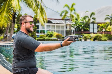 Sakallı adam uçuş için drone başlattı, hangi ile fotoğraf ve video çekimi alabilir, yüzme havuzu ve palmiye ağaçları yakınında, yan görünüm.