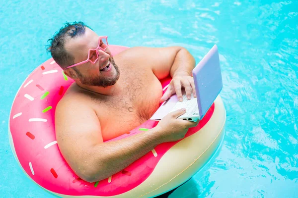 laughing Fat funny man in pink inflatable circle in pink glasses works on a laptop in a swimming pool