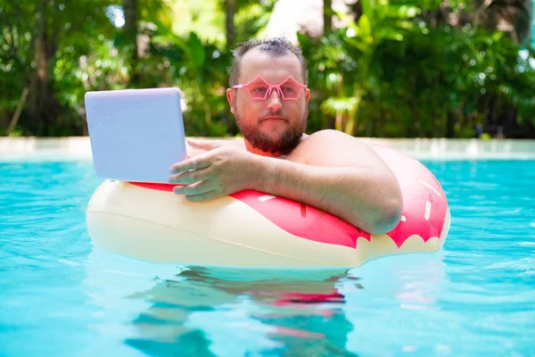Fat funny man in pink inflatable circle in pink glasses works on a laptop in a swimming pool