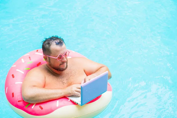 Серйозна товста смішна людина в рожевому надувному колі в рожевих окулярах працює на ноутбуці в басейні — стокове фото