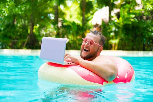 Сміється Товстий смішний чоловік в рожевому надувному колі в рожевих окулярах працює на ноутбуці в басейні — стокове фото