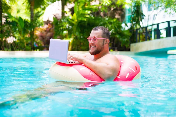 smiling Fat funny man in pink inflatable circle in pink glasses works on a laptop in a swimming pool