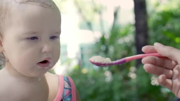 Una ragazza di un anno mangia un cucchiaio di porridge in natura vicino a palme, tropici, rallentatore — Video Stock