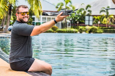 Sakallı adam uçuş için drone başlattı, hangi ile fotoğraf ve video çekimi alabilir, yüzme havuzu ve palmiye ağaçları yakınında, yan görünüm.