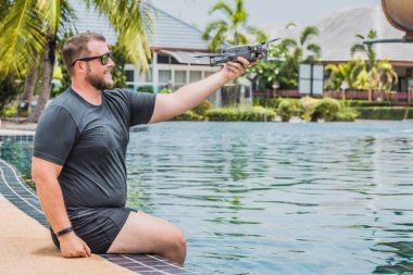 Sakallı adam uçuş için drone başlattı, hangi ile fotoğraf ve video çekimi alabilir, yüzme havuzu ve palmiye ağaçları yakınında, yan görünüm.