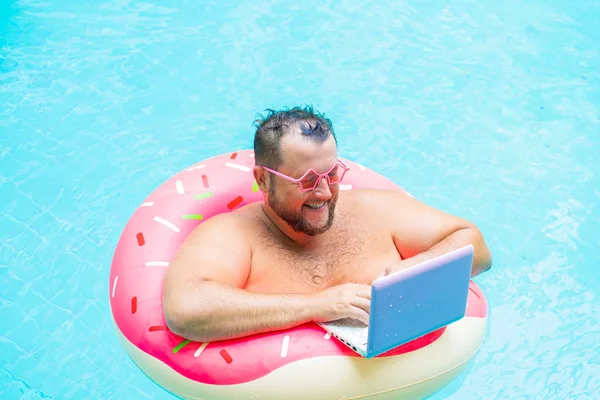Сміється Товстий смішний чоловік в рожевому надувному колі в рожевих окулярах працює на ноутбуці в басейні — стокове фото