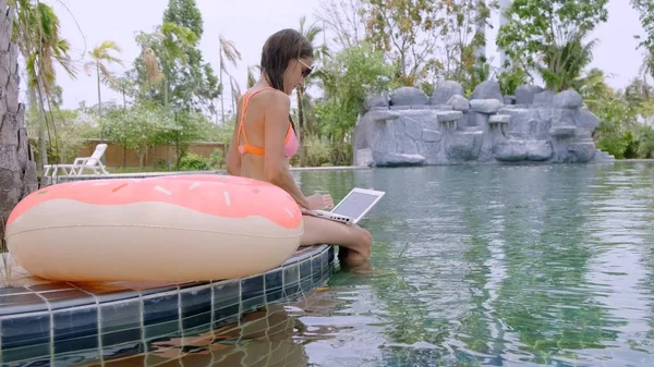 Young female freelancer sitting with laptop near pool. Busy during the holidays. The concept of remote work.