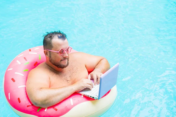 serious Fat funny man in pink inflatable circle in pink glasses works on a laptop in a swimming pool