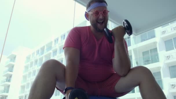 Funny fat hane i rosa glas och i en rosa t-shirt är engagerad med hantlar på en fit Ball i gymmet som skildrar en flicka — Stockvideo