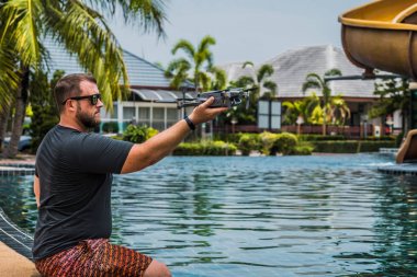 Sakallı adam uçuş için drone başlattı, hangi ile fotoğraf ve video çekimi alabilir, yüzme havuzu ve palmiye ağaçları yakınında, yan görünüm.