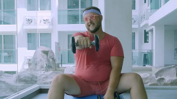 Funny grasa masculina en gafas de color rosa y en una camiseta rosa se dedica con mancuernas en una bola en forma en el gimnasio que representa a una chica —  Fotos de Stock