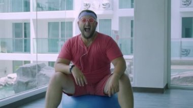 Pembe gözlük ve pembe bir t-shirt komik şişman erkek bir kız tasvir spor salonunda uygun bir top üzerinde meşgul. Harika bir adam spor salonunda top müzik ve dans dinler.