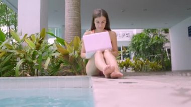 kadın Freelancer havuz tarafından oturur ve bir minicomputer üzerinde çalışır, kız suya onun laptop düşer. Tatil sırasında meşgul. Uzak çalışma kavramı. seçici odak