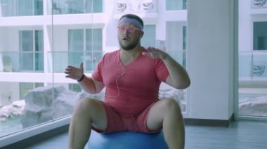 Pembe gözlük ve pembe bir t-shirt komik şişman erkek bir kız tasvir spor salonunda uygun bir top üzerinde meşgul. Harika bir adam spor salonunda top müzik ve dans dinler.
