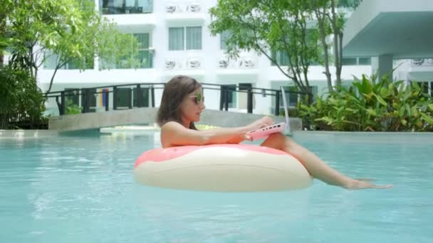 Joven freelancer sentada con un portátil en un círculo inflable en la piscina. Ocupado durante las vacaciones. Concepto de trabajo remoto. enfoque selectivo — Vídeos de Stock