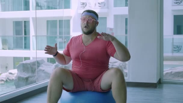 Funny grasa masculina en gafas de color rosa y en una camiseta rosa se dedica a una bola en forma en el gimnasio que representa a una chica. Un hombre maravilloso escucha música y baila en la pelota en el gimnasio . — Vídeos de Stock