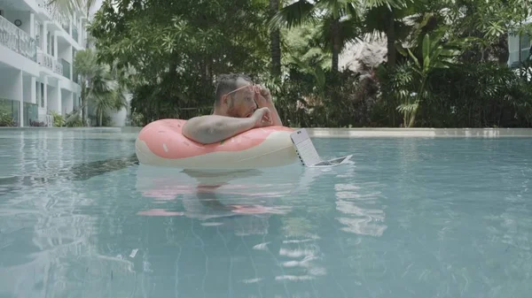 Dikke grappige man in roze opblaasbare cirkel in roze bril werkt op een laptop in een zwembad. De man is verrast en daalt de laptop in het water in het zwembad. — Stockfoto