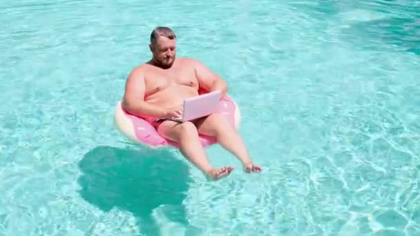 Мужчина с надувным кольцом в воде в бассейне работает за компьютером. Смешной толстяк в круге плавания с розовым ноутбуком. внештатная концепция — стоковое видео