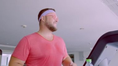 Pembe gözlük ve pembe bir t-shirt komik şişman erkek bir kız tasvir spor salonunda bir koşu bandı üzerinde meşgul. 4k