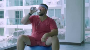 Pembe gözlük ve pembe bir t-shirt komik şişman erkek bir kız tasvir spor salonunda uygun bir top üzerinde meşgul. Eksantrik adam spor salonunda topu bir şişe su içiyor.