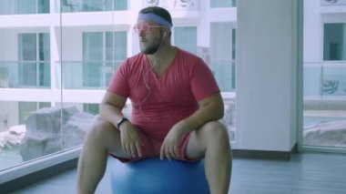 Pembe gözlük ve pembe bir t-shirt komik şişman erkek bir kız tasvir spor salonunda uygun bir top üzerinde meşgul. Harika bir adam spor salonunda top müzik ve dans dinler.