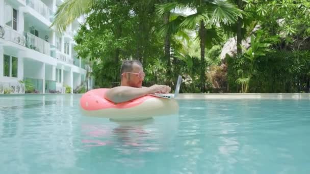Dikke grappige man in roze opblaasbare cirkel in roze bril werkt op een laptop in een zwembad — Stockvideo