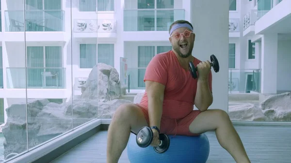 Funny fat male in pink glasses and in a pink t-shirt is engaged with dumbbells on a fit ball in the gym depicting a girl