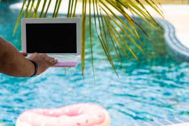 Erkek bilgisayarında çalışıyor, havuzun yanında oturuyor. Havuzda klavyede erkek eller