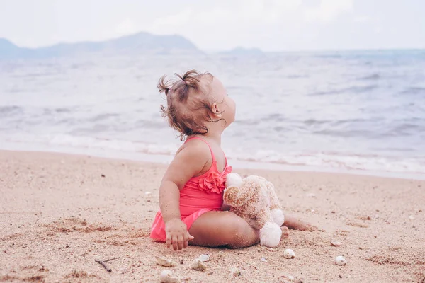 Задумчивая маленькая девочка обнимает плюшевого мишку и смотрит в сторону, сидя на берегу моря . — стоковое фото