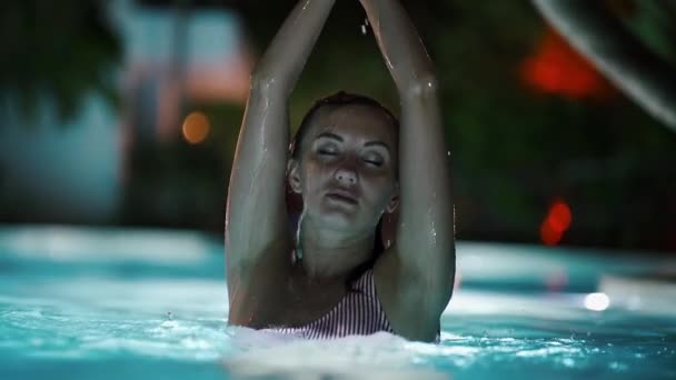 Сексуальная девушка отдыхает в бассейне ночью. самка поднимает руки вверх, и вода брызгает — стоковое видео