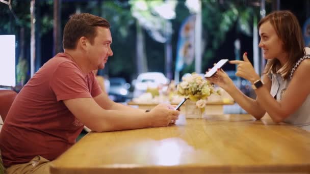 Couple amoureux homme et femme sont assis dans un café à la table sur un rendez-vous le soir. Femme montre à l'homme quelque chose au téléphone, homme sourit — Video