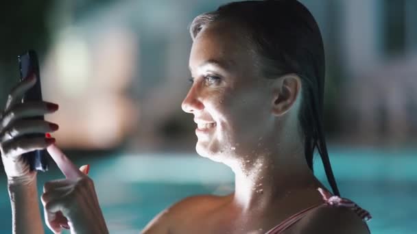 Mulher usando aplicativo de telefone celular em férias na piscina à noite em férias de viagem. Menina usando aplicativo smartphone olhando para a tela sorrindo feliz. Tela de luz no rosto — Vídeo de Stock