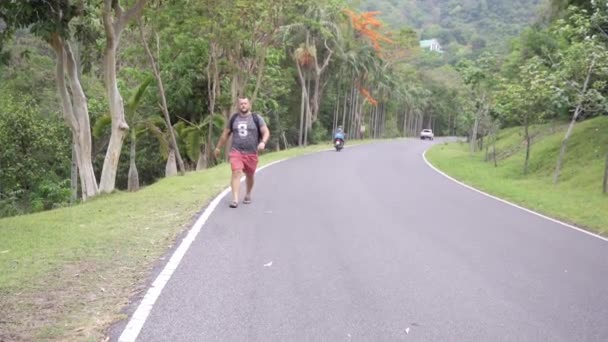 Bonita floresta tropical com um jovem viajante com uma mochila na estrada para a floresta da Tailândia. Um homem caminha em uma estrada tropical e desfruta das vistas da natureza — Vídeo de Stock