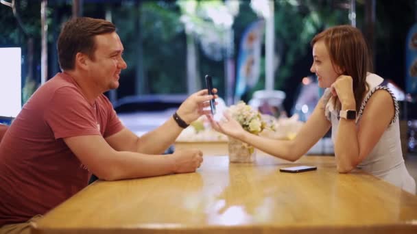 Couple in love man and woman are sitting in a cafe at the table on a date in the evening. Man shows woman something on the phone, girl smiles — Stock Video