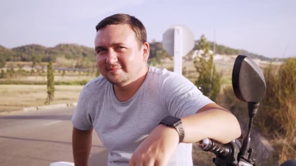 Retrato de um jovem sentado em uma motocicleta perto de uma estrada entre campos e colinas, close-up — Vídeo de Stock