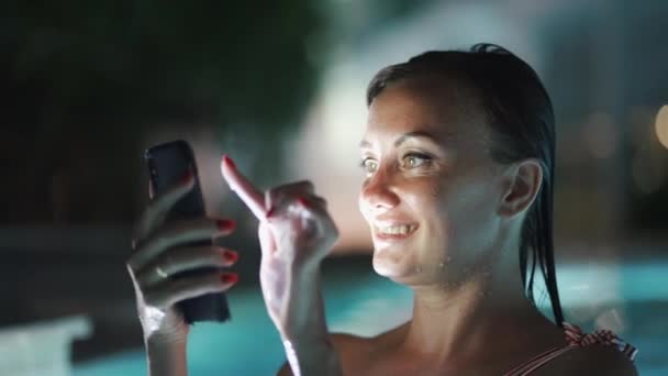 Donna che utilizza l'app per cellulare in vacanza in piscina di notte durante le vacanze di viaggio. Ragazza utilizzando app smartphone guardando lo schermo sorridente felice. Luce dello schermo sul viso — Video Stock