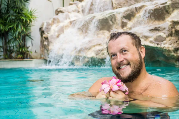 Lachende bebaarde man houdt bloemen in het zwembad. Tropische bloemen Frangipani Plumeria, Leelawadee drijvend in het water. Spabad. Vrede en rust. Spa concept. — Stockfoto