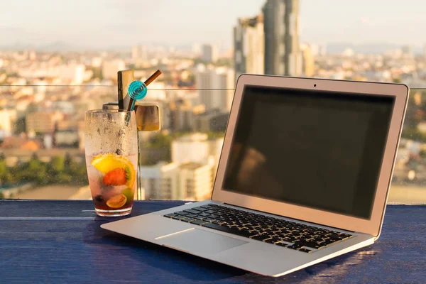 Ноутбук і келих літнього коктейлю на дерев'яному столі, на яскравому красивому панорамному тлі міста. макет — стокове фото